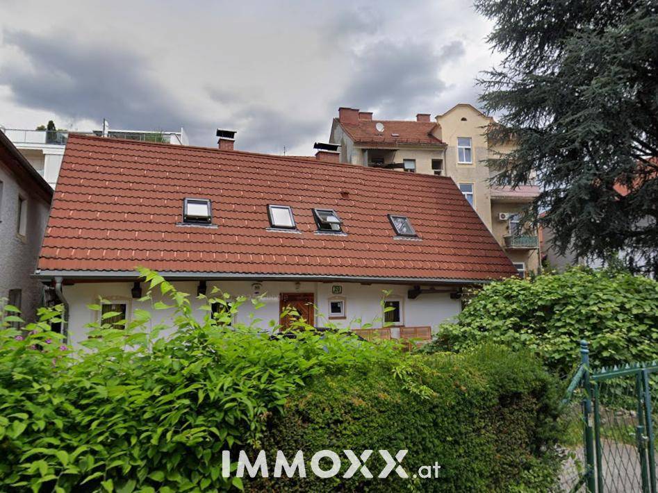 Ansicht des Hauses mit rotem Ziegeldach, Dachfenstern und einer hohen grünen Hecke im Vordergrund.