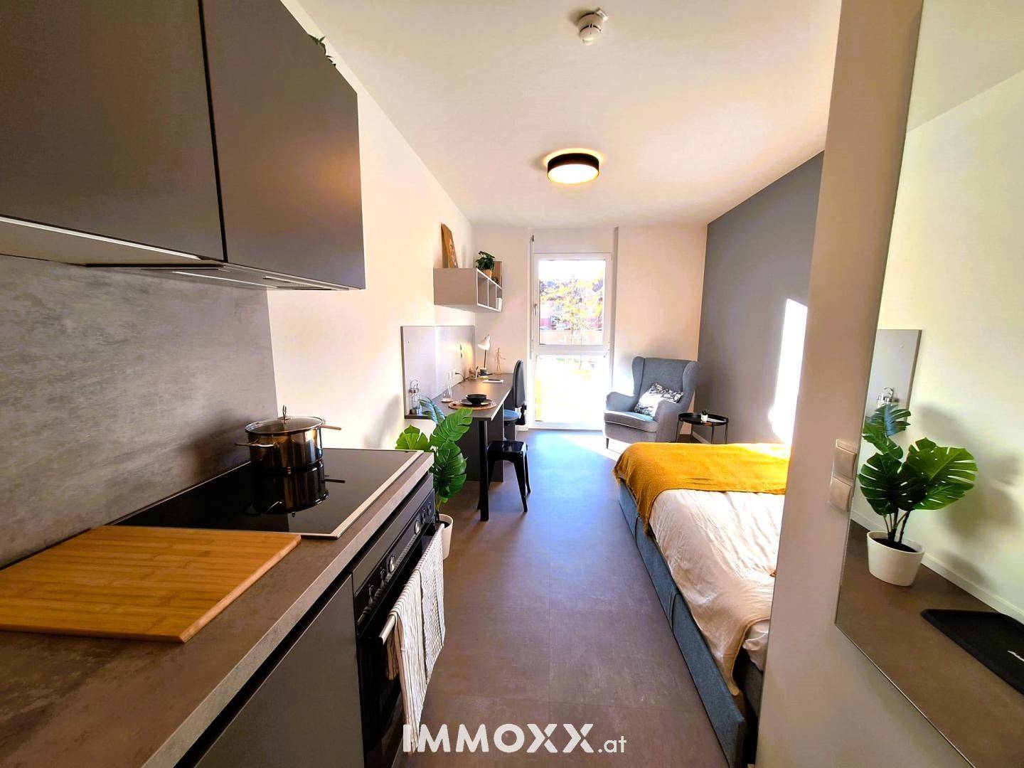 Helles Studio-Apartment mit Küchenzeile, Bett, Sessel und Blick nach draußen.