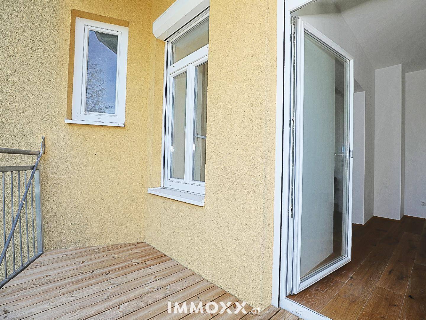Balkon mit Holzbelag und Zugang zum Innenraum, umgeben von einer gelben Fassade.