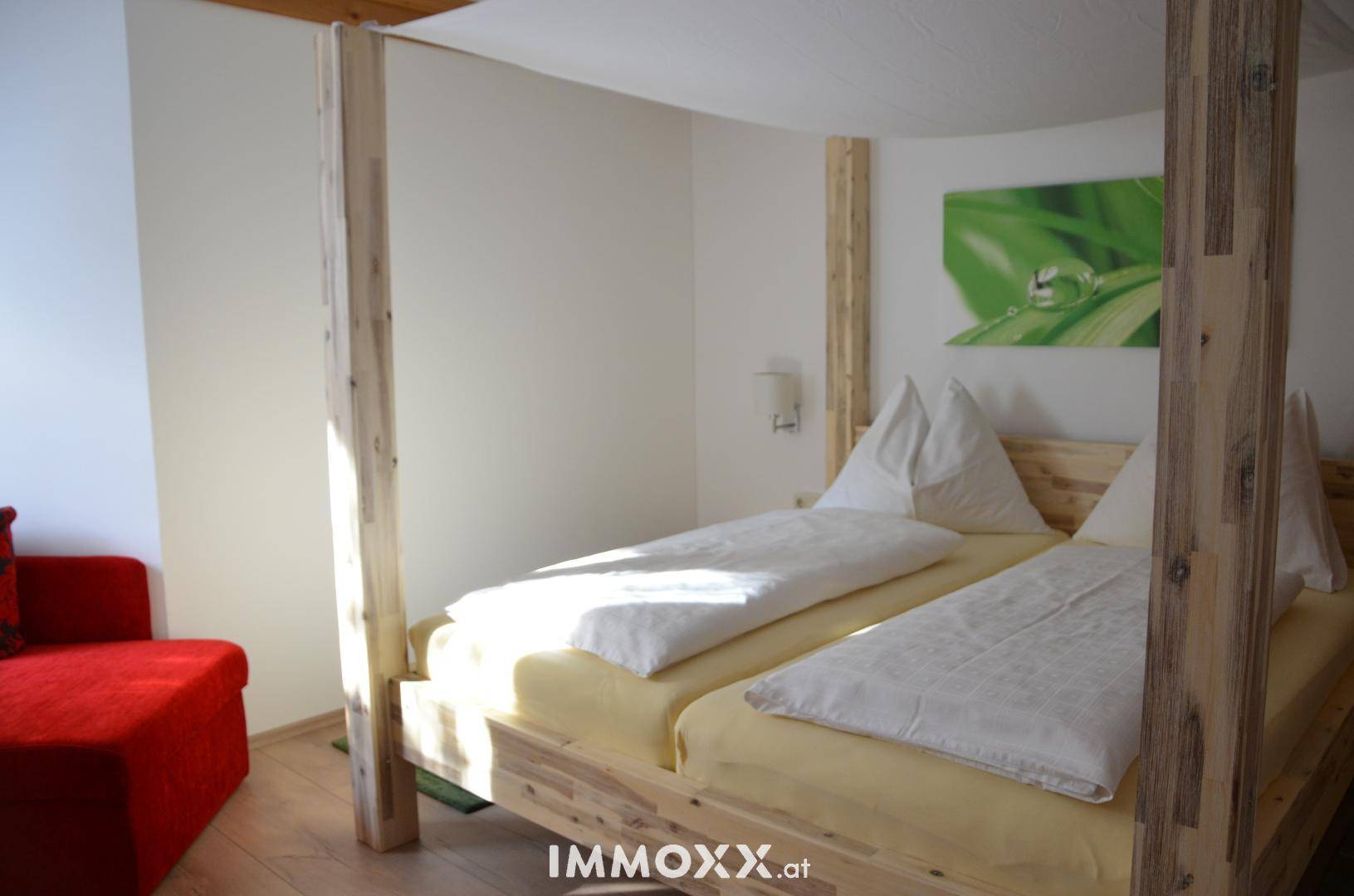 Helles Schlafzimmer mit einem Himmelbett aus hellem Holz und einem bequemen roten Sessel.