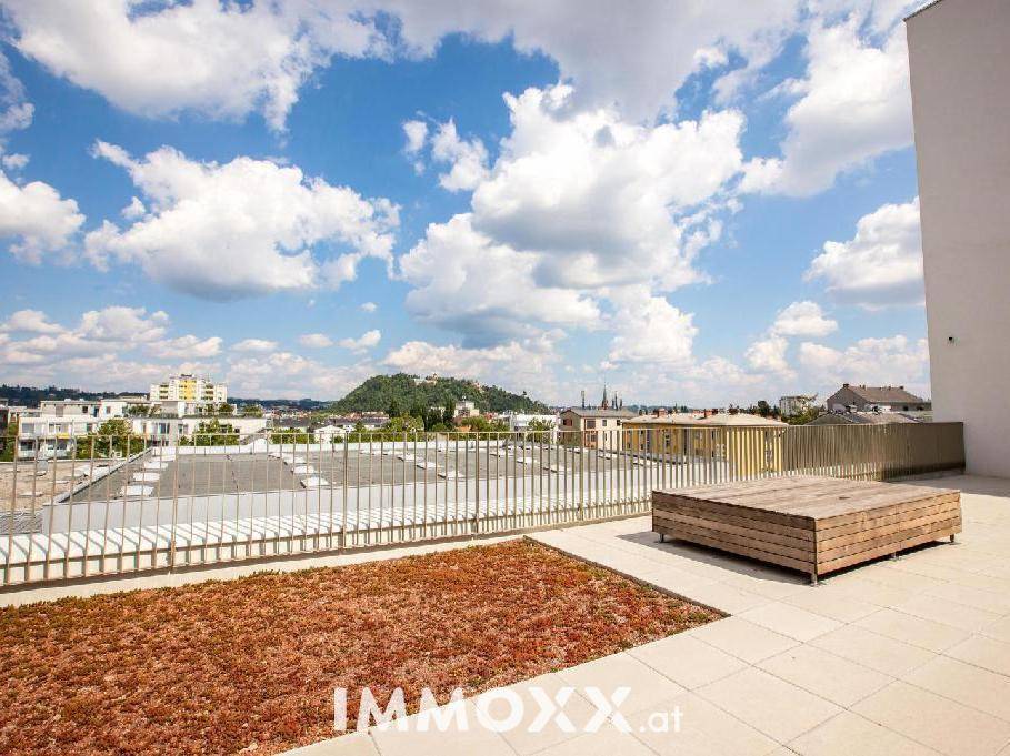Dachterrasse mit Stadtblick, Sitzgelegenheit und begrüntem Bereich unter blauem Himmel.