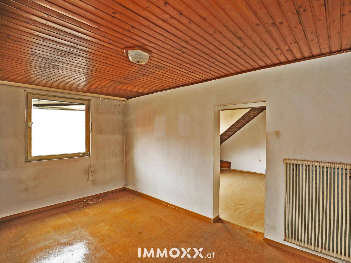 Heller Raum mit Holzdecke und Fenster, bietet Zugang zu einem weiteren Bereich mit Treppe.