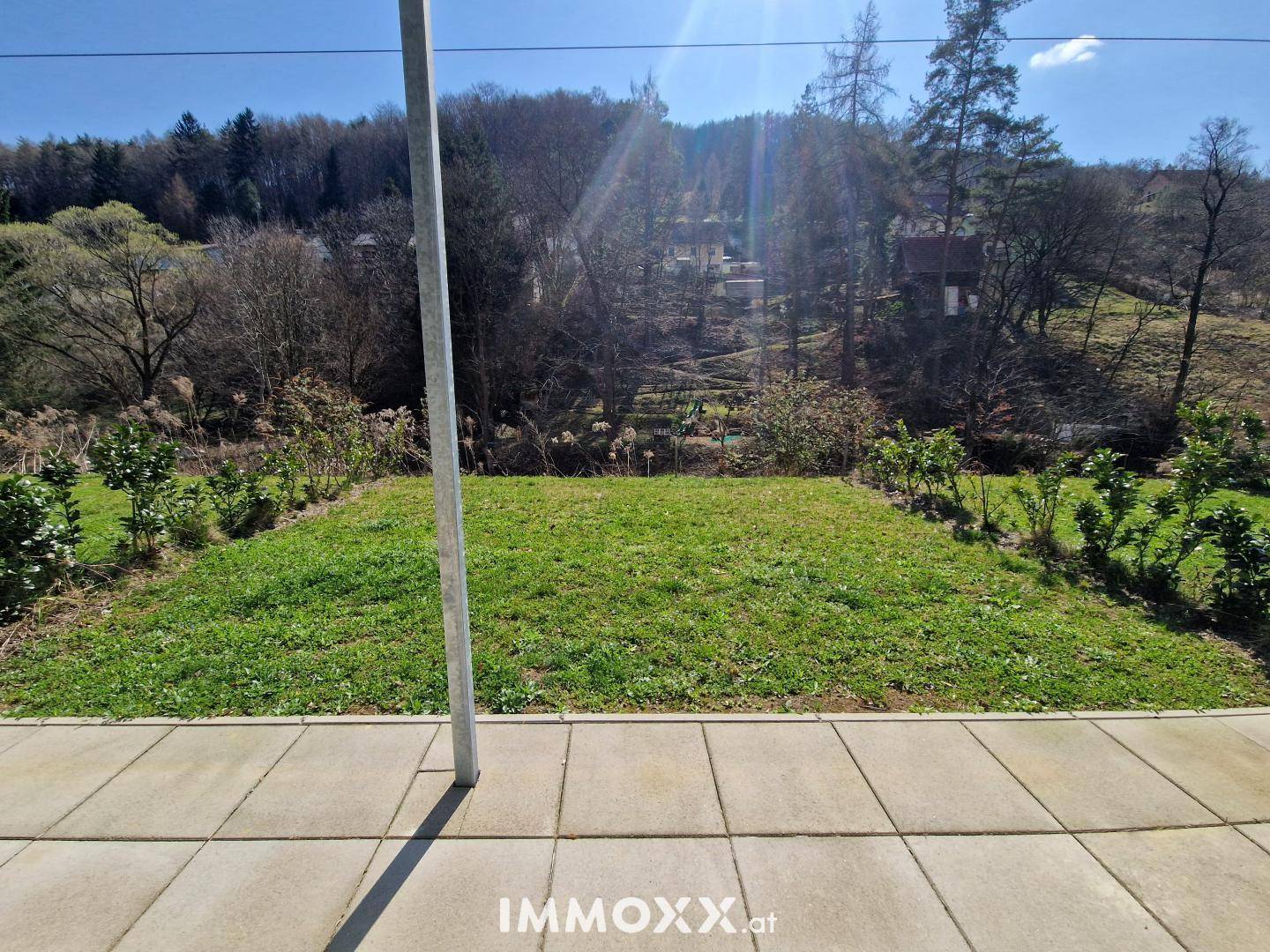 Gepflegte Rasenfläche mit Bäumen im Hintergrund, gesehen von einer gepflasterten Terrasse.