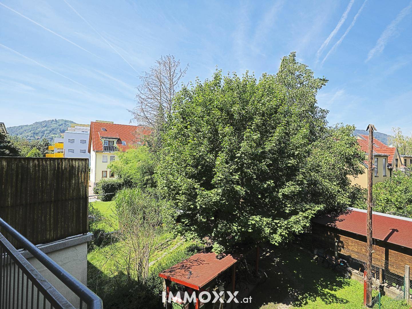 Balkonblick ins Grüne mit Bäumen und Dächern der umliegenden Häuser unter blauem Himmel.
