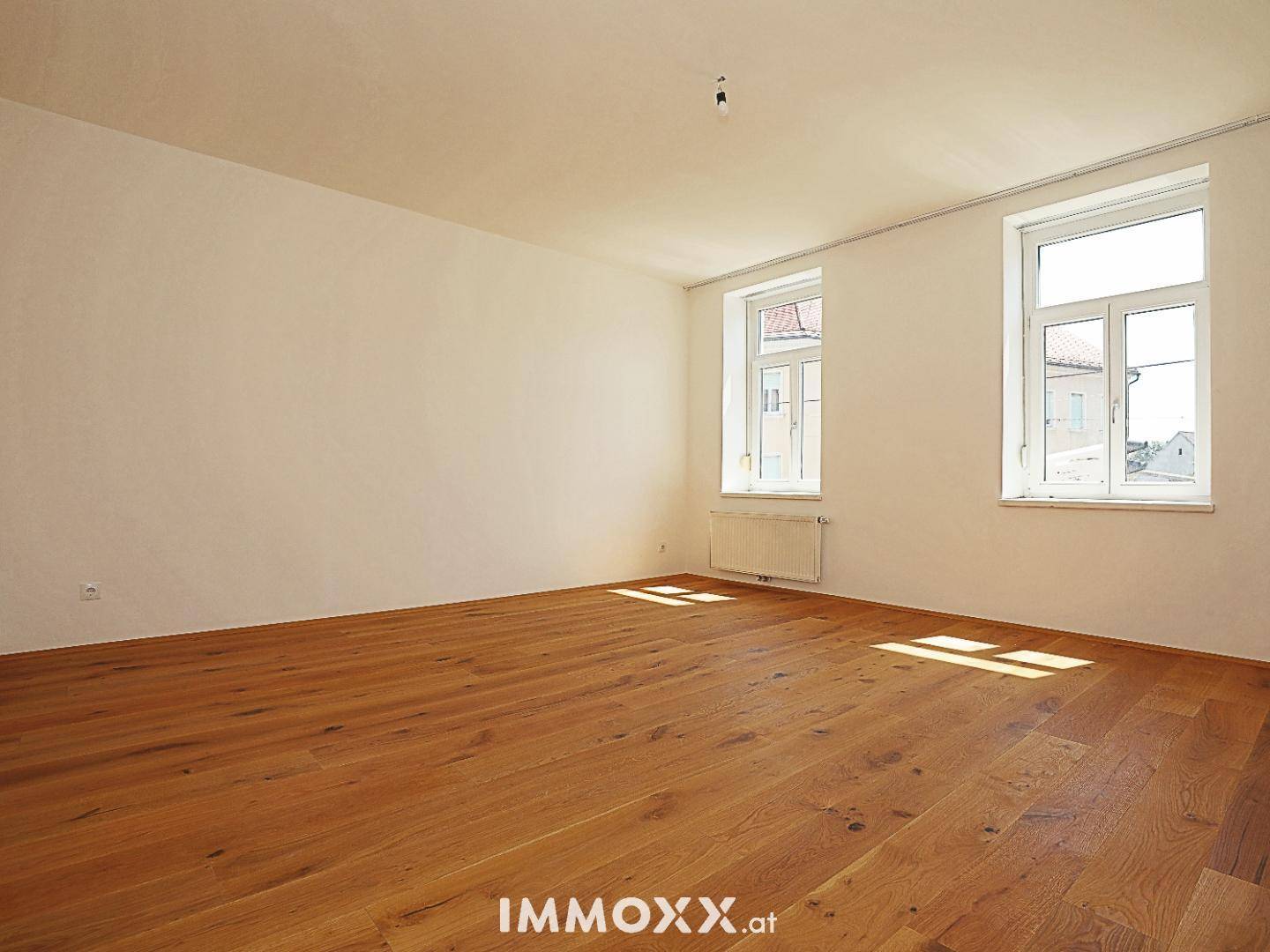 Heller Raum mit Holzboden und zwei Fenstern, die einen Blick auf die Umgebung ermöglichen.