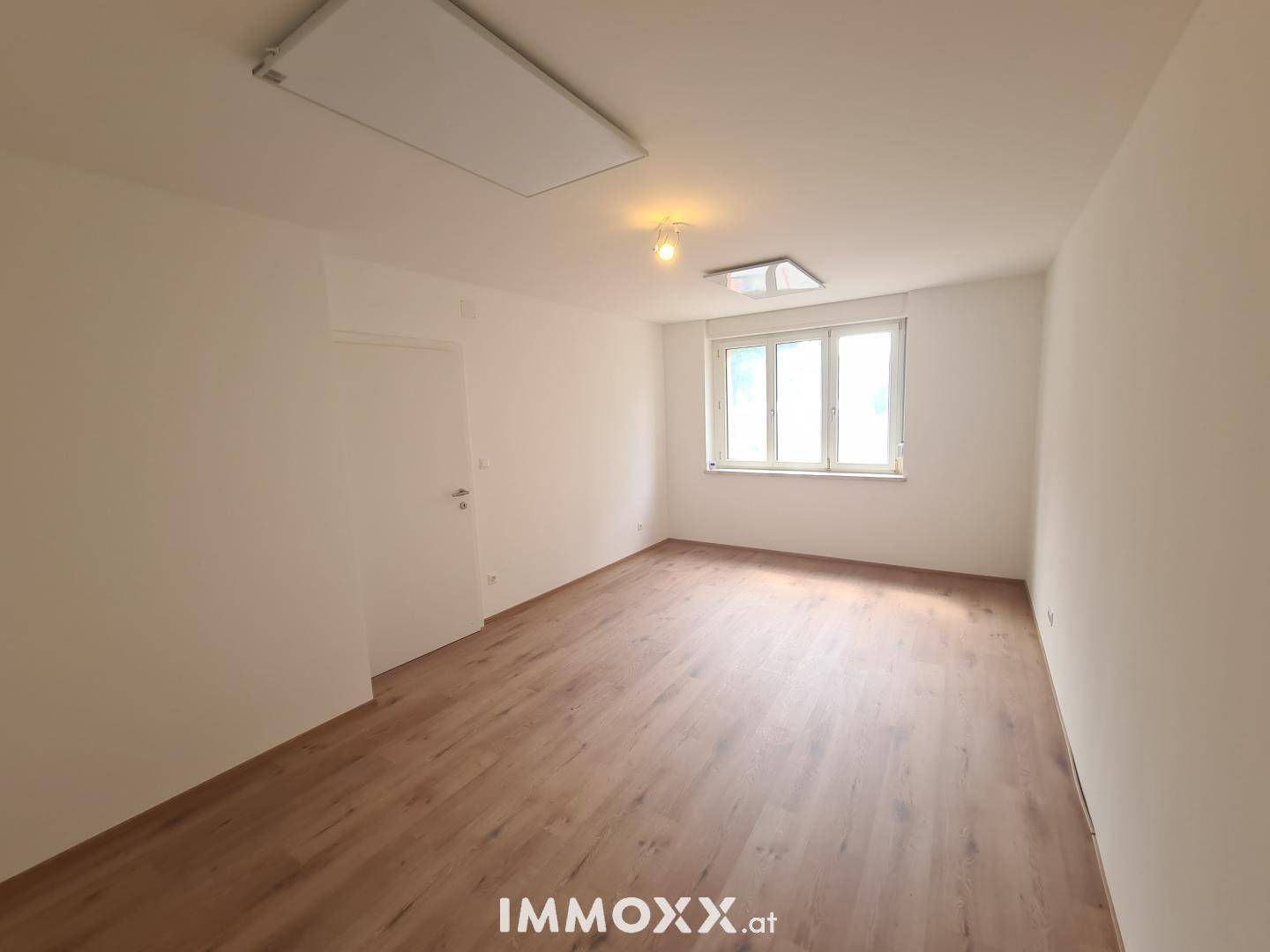 Leeres Zimmer mit Laminatboden, weißen Wänden und einem Fenster, das viel Tageslicht hereinlässt.