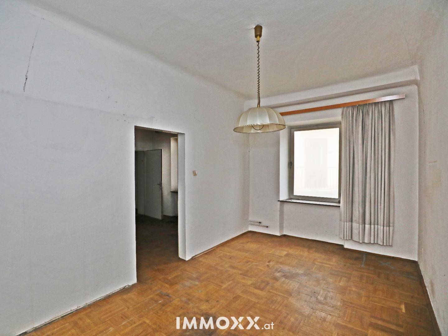 Leeres Zimmer mit abgenutztem Parkettboden, weißen Wänden und einem Fenster mit Vorhängen, das Renovierungsbedarf zeigt.