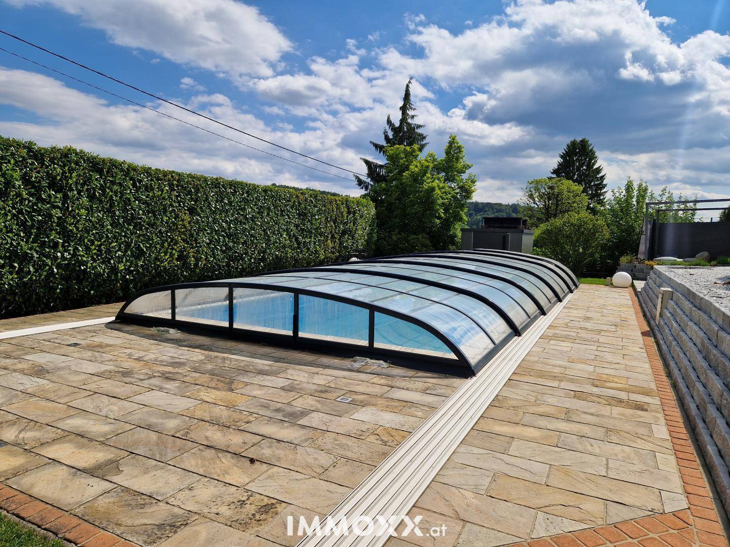 Überdachter Swimmingpool im Garten, umgeben von einer gepflasterten Terrasse und einer hohen Hecke.