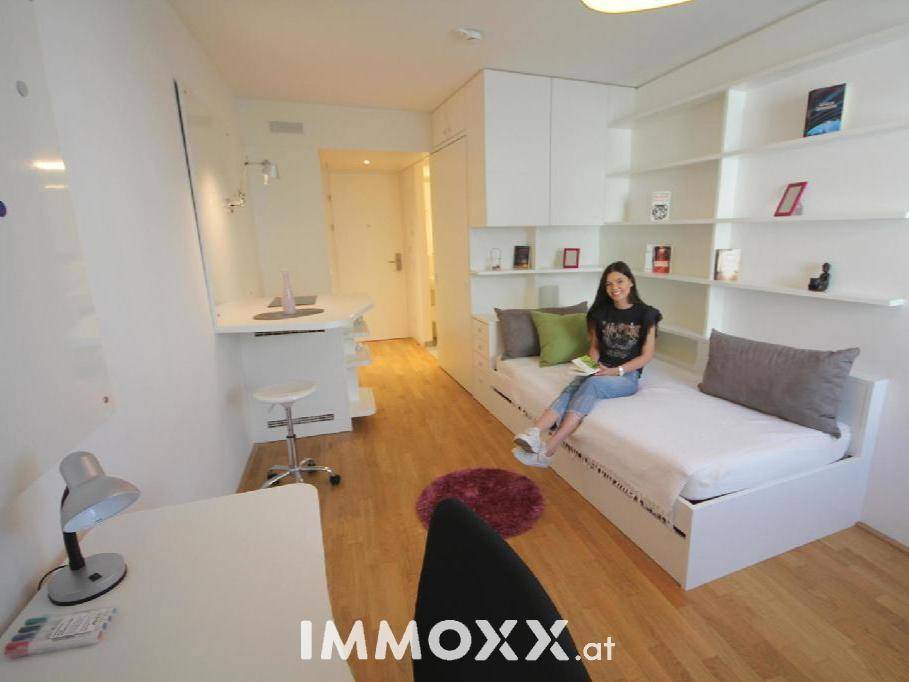 Helles Studio-Apartment mit Bett, Schreibtisch und integrierten Regalen für effizientes Wohnen.