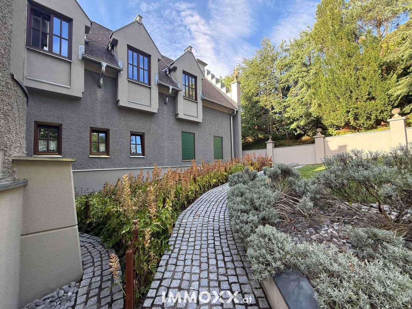 Gepflasterter Gartenweg entlang des Gebäudes, umgeben von gepflegten Pflanzen und Sträuchern.