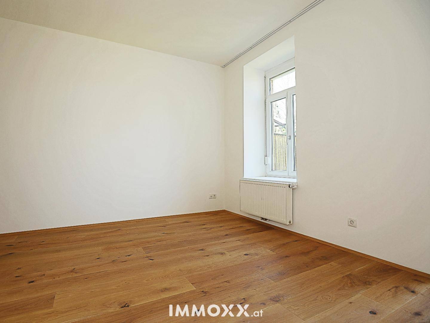 Helles, leeres Zimmer mit Holzboden und einem Fenster, das viel Tageslicht hereinlässt.