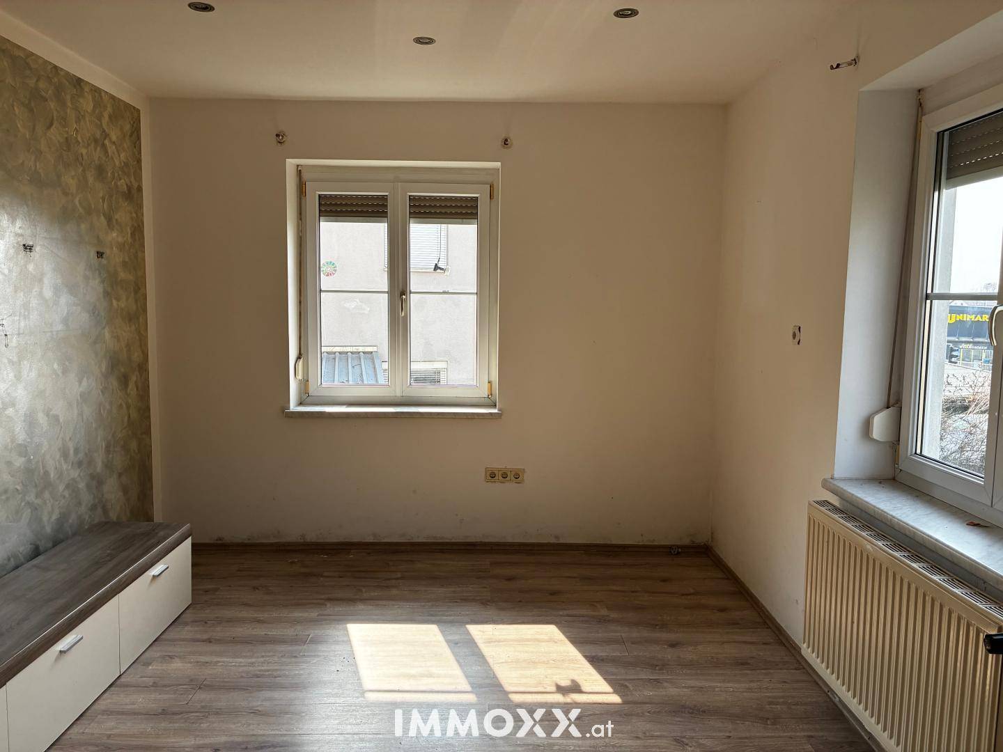 Helles Zimmer mit Laminatboden, einem Fenster und einer Wandbank.