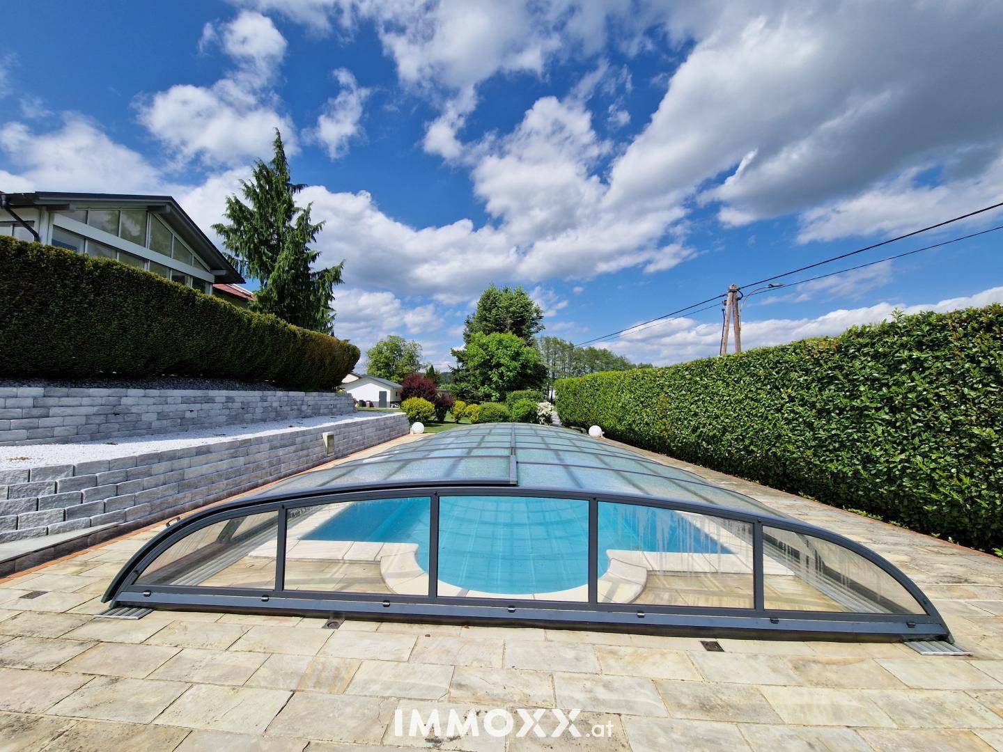 Großer Swimmingpool mit Schiebeüberdachung, umgeben von einer gepflasterten Fläche und grünen Hecken.