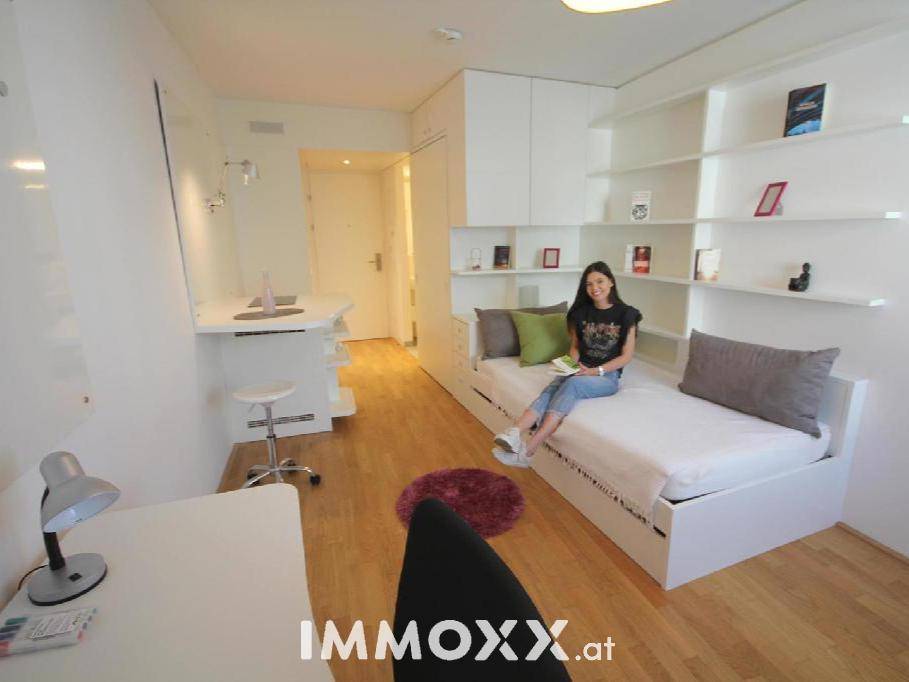 Helles Studio-Apartment mit Bett, Schreibtisch und integrierten Regalen für effizientes Wohnen.