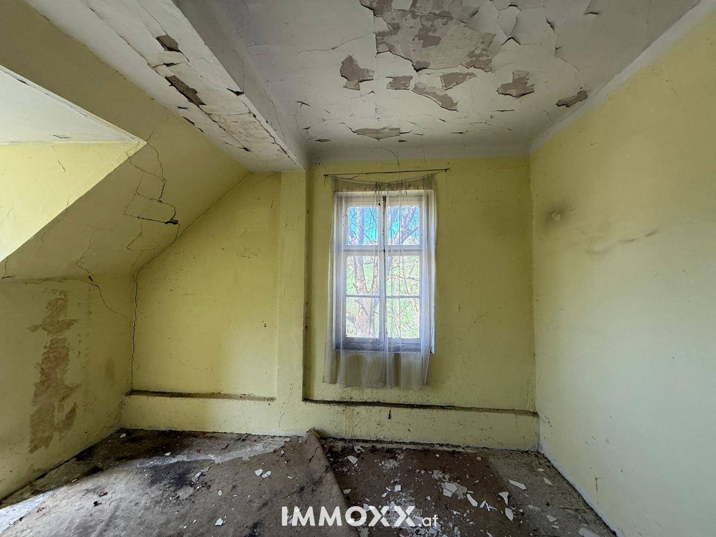 Ein kleines Zimmer mit gelben Wänden, einem Fenster und stark abblätternder Farbe an Decke und Wänden.