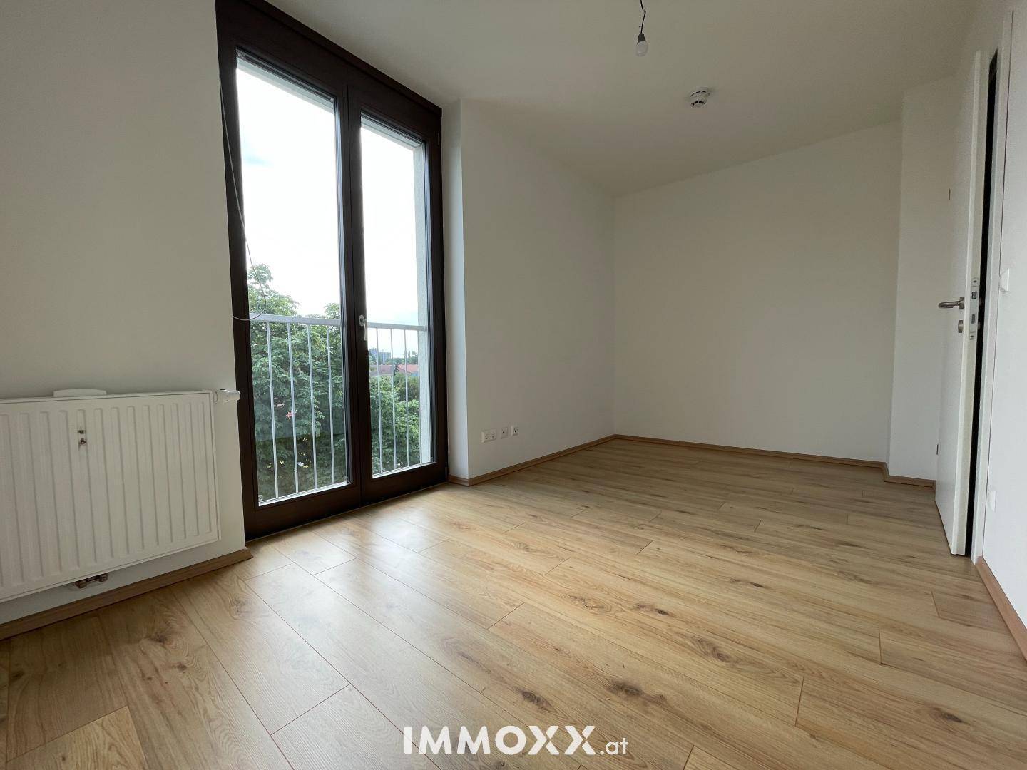 Helles, leeres Zimmer mit Holzboden, großen Fenstern und Zugang zu einem französischen Balkon.