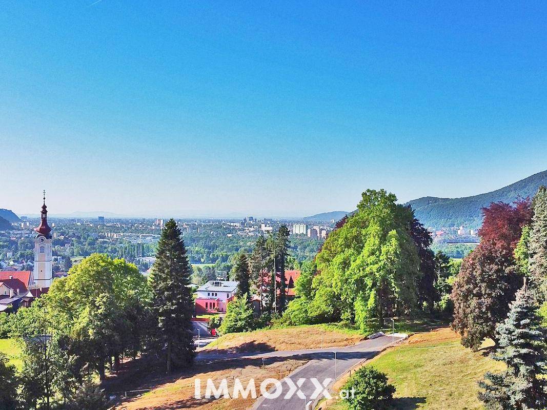 Weitläufiger Panoramablick über eine Stadt mit Kirchturm, umgeben von grünen Hügeln und Bergen unter blauem Himmel.