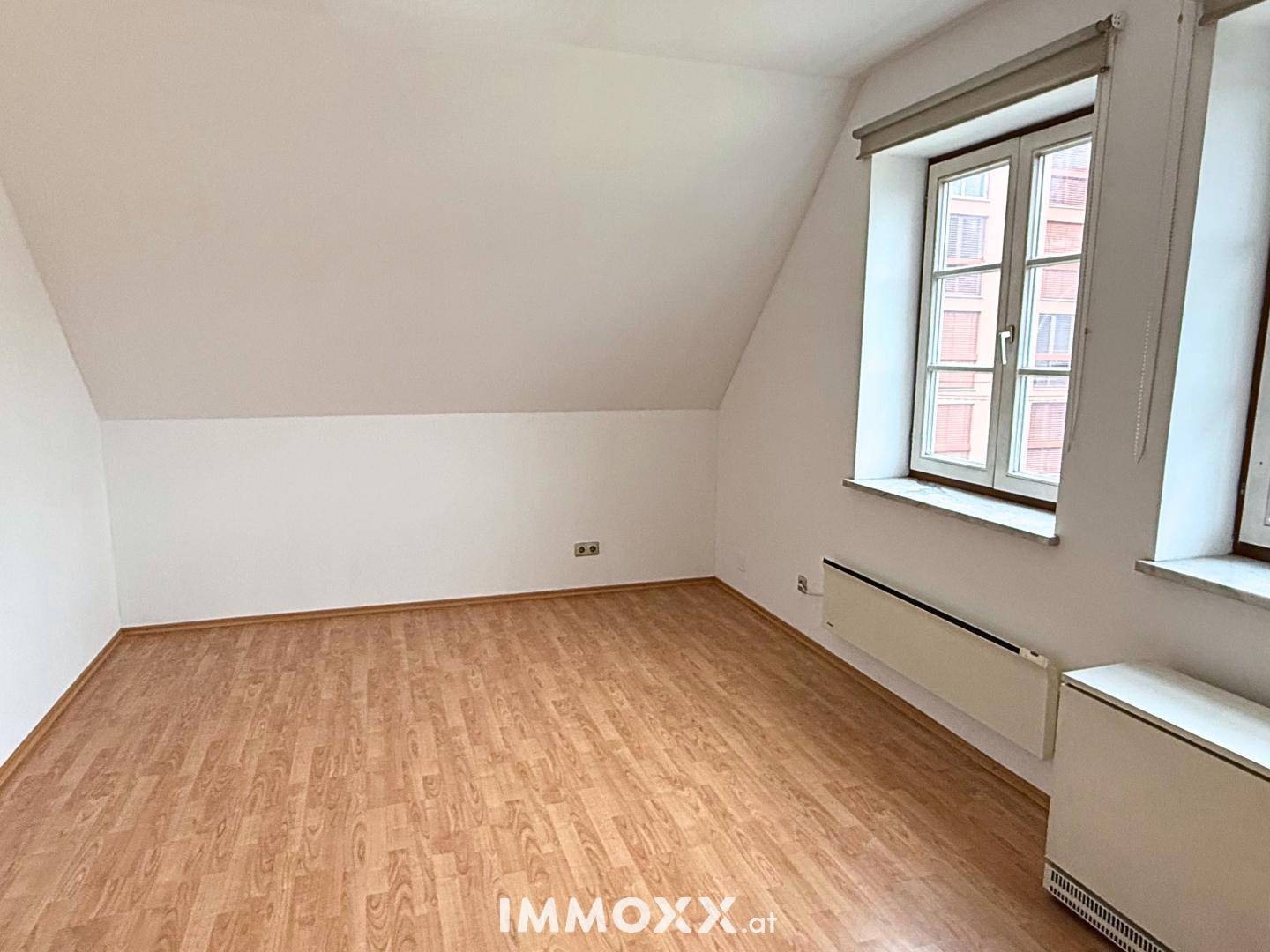 Leerer Raum mit Laminatboden, weißen Wänden und einem Fenster unter der Dachschräge.