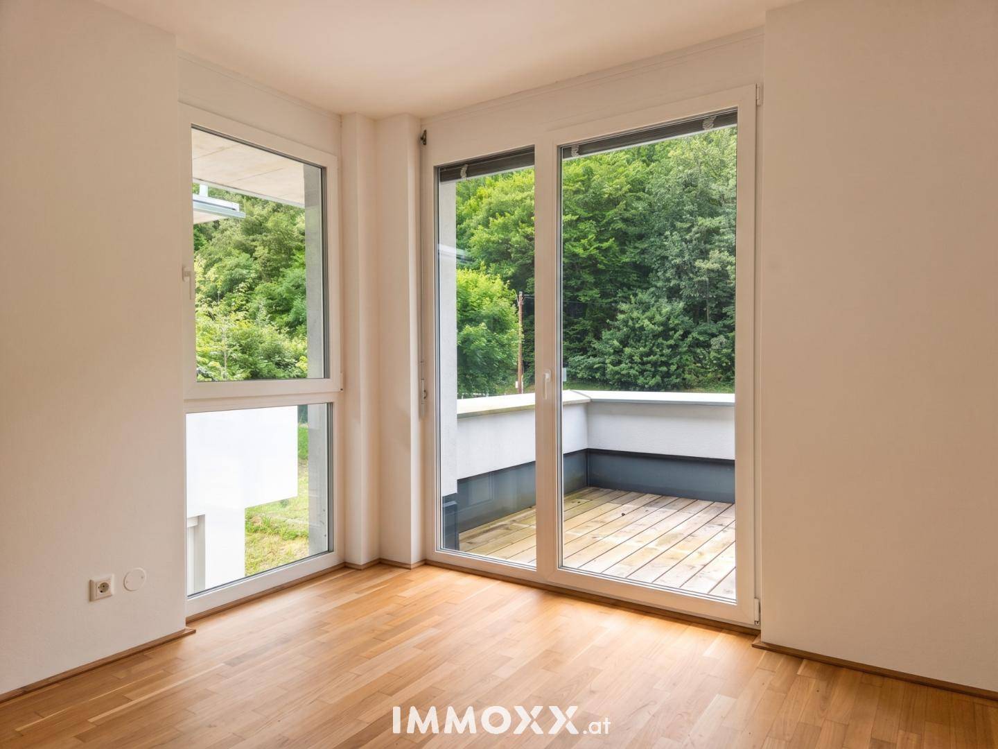 Heller Raum mit Holzboden und großen Fenstern, die Zugang zu einem Balkon bieten und Blick ins Grüne.