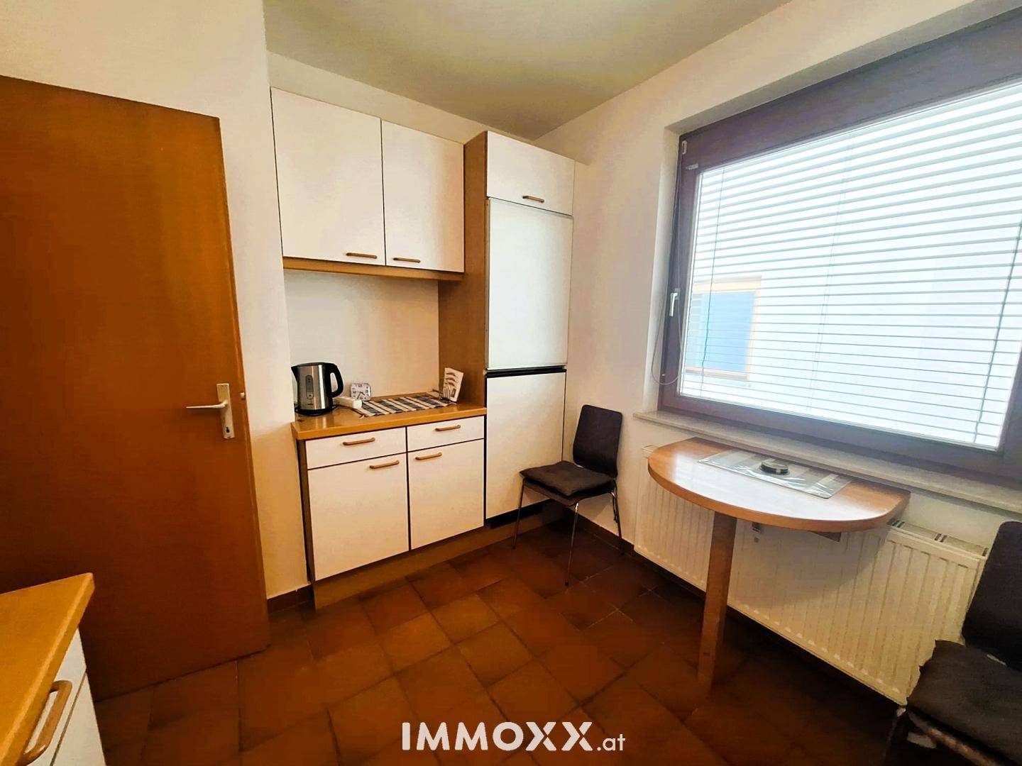 Kleine Küche mit weißen Schränken, Kühlschrank und einem Essbereich am Fenster.