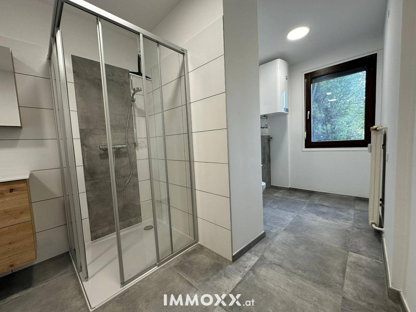 Modernes Badezimmer mit Duschkabine, grauen Fliesen und einem Fenster für Tageslicht.