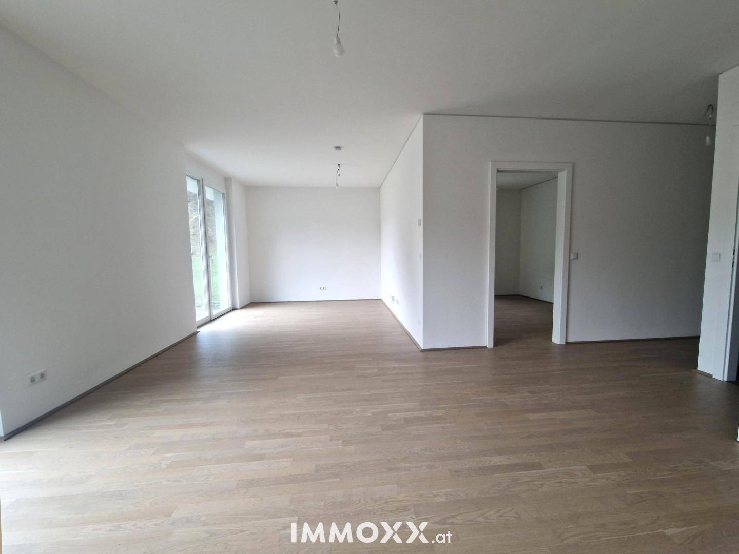 Geräumiges, leeres Wohnzimmer mit hellem Parkettboden und Zugang zu einem Außenbereich.
