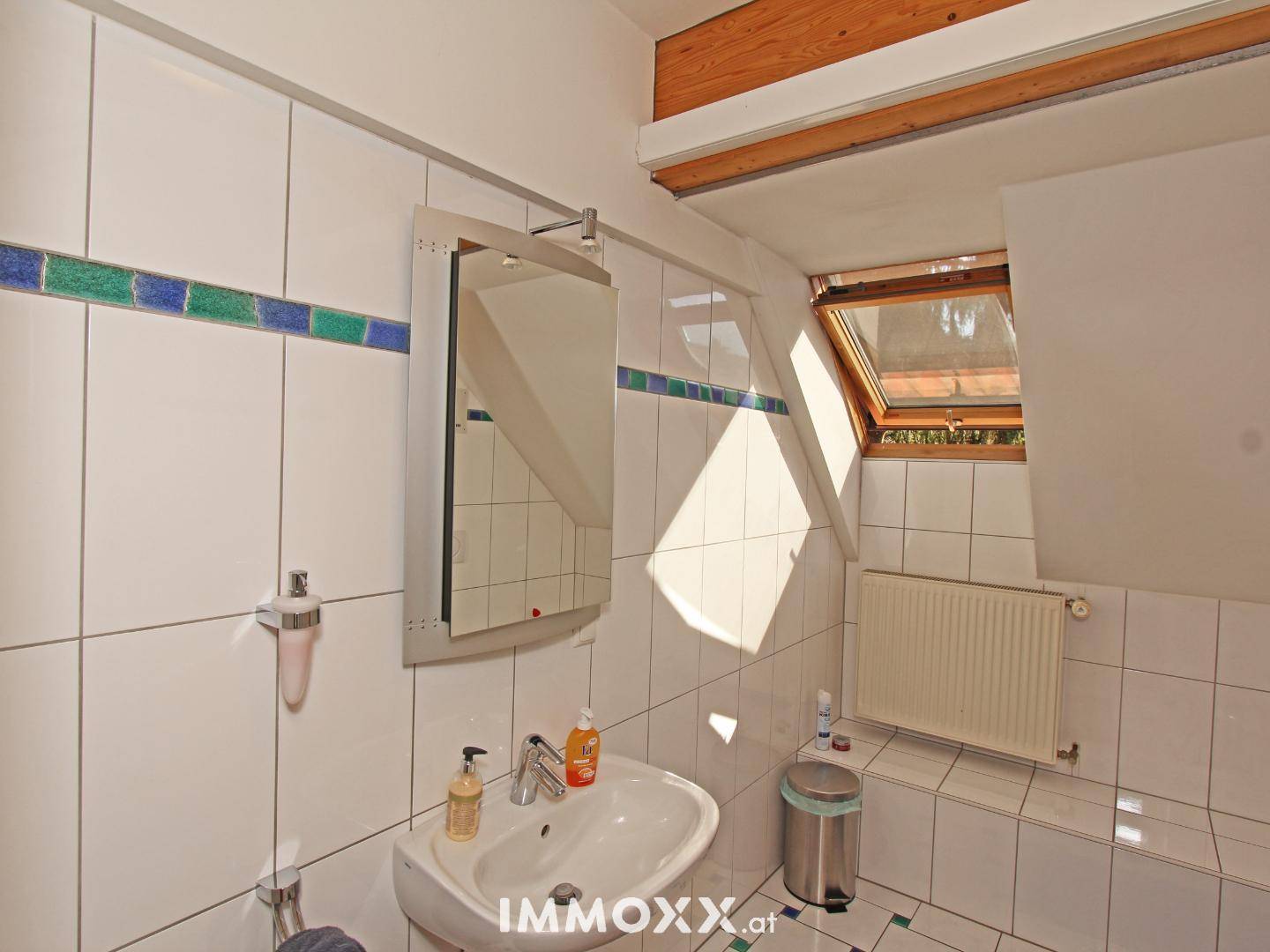 Badezimmer mit weißen Fliesen, Waschbecken, Spiegel und einem Dachfenster für natürliches Licht.