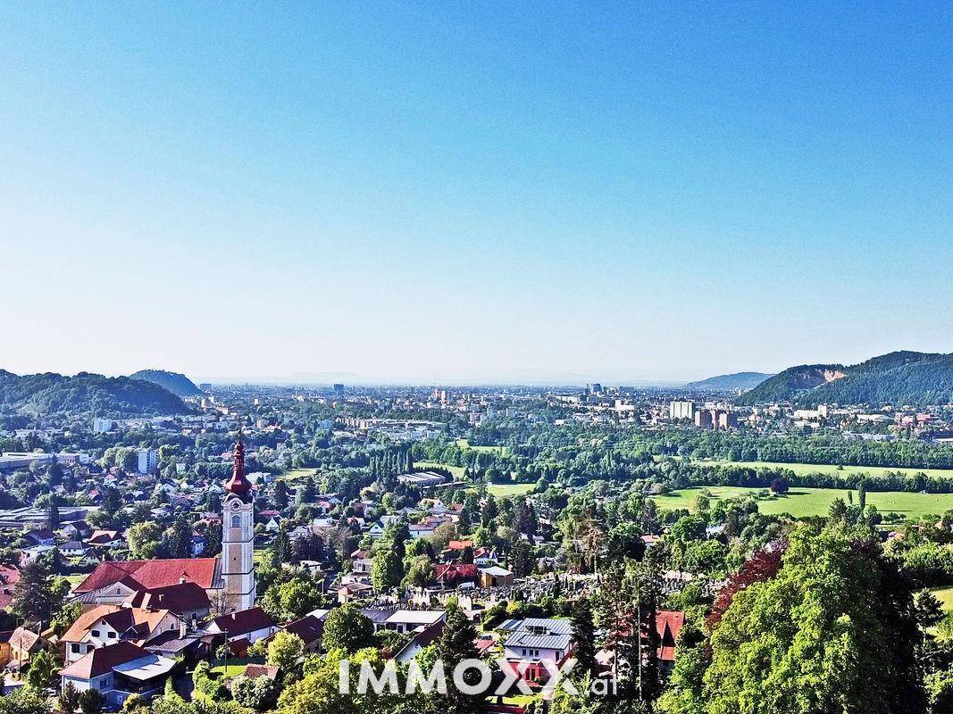 Weitläufiger Panoramablick über eine Stadt mit Kirchturm, umgeben von grünen Hügeln und Bergen.