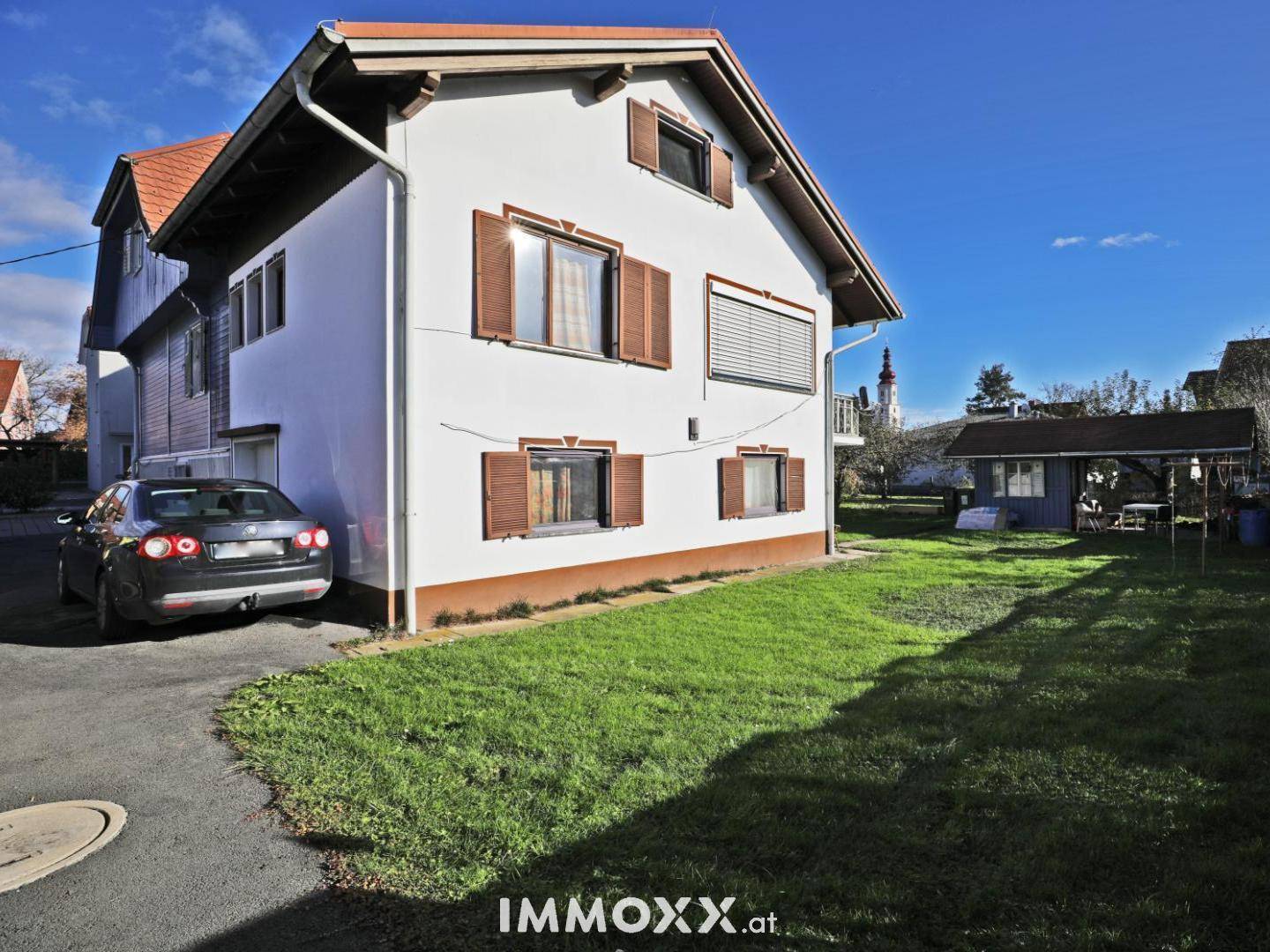 Seitenansicht des weißen Hauses mit Rasenfläche, Auffahrt und einem Carport im Hintergrund.