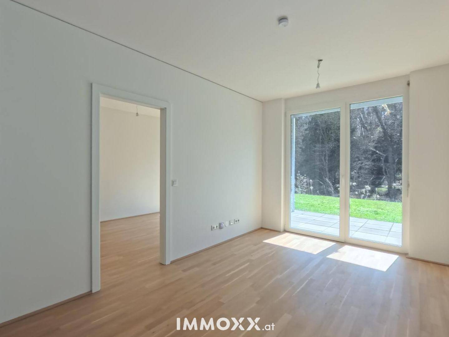 Leerer Wohnraum mit hellem Parkettboden und großen Fenstern zum Garten.