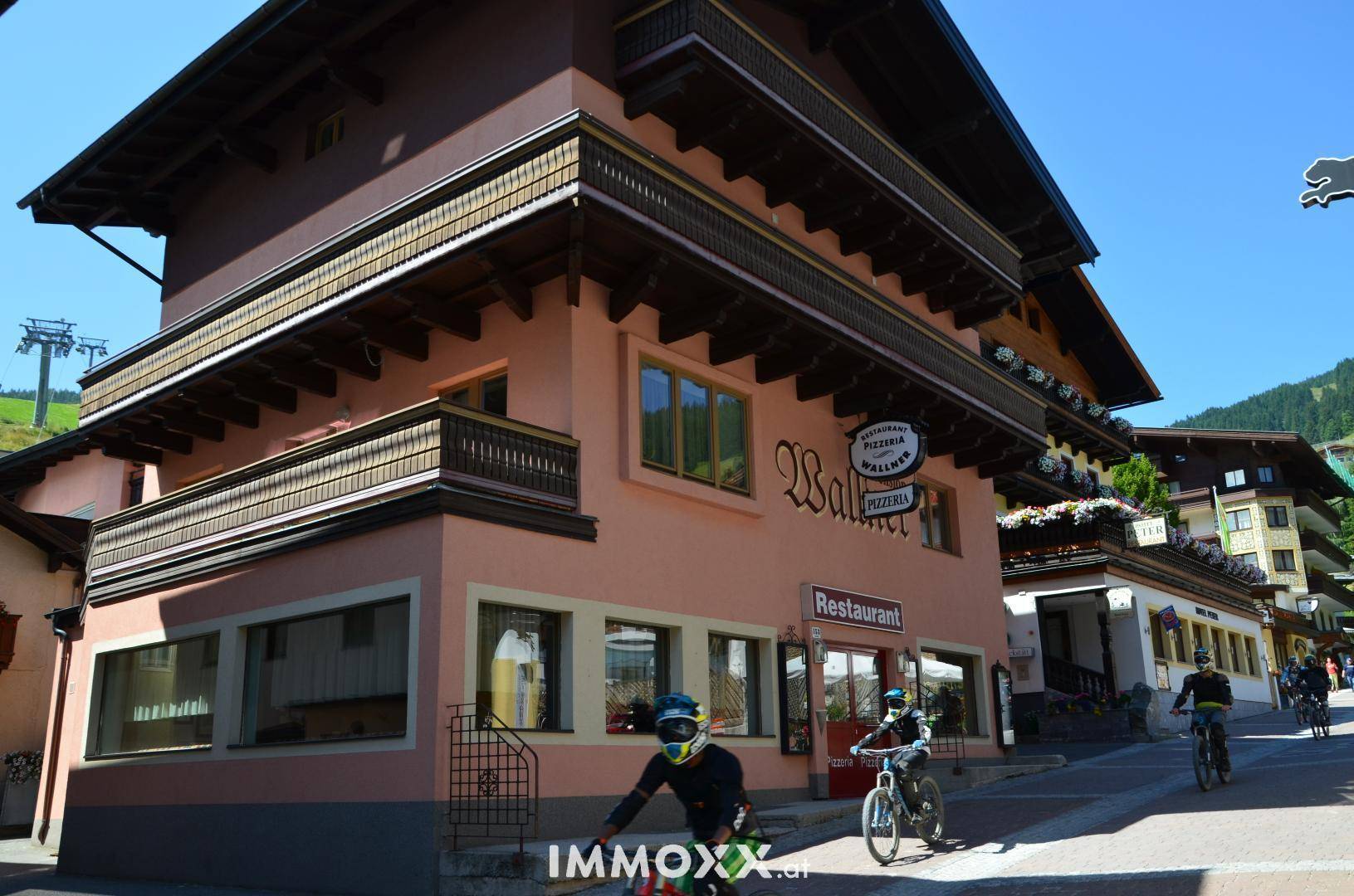Traditionelles Gebäude mit rosa Fassade, Holzbalkonen und einem Restaurant im Erdgeschoss an einem sonnigen Tag.
