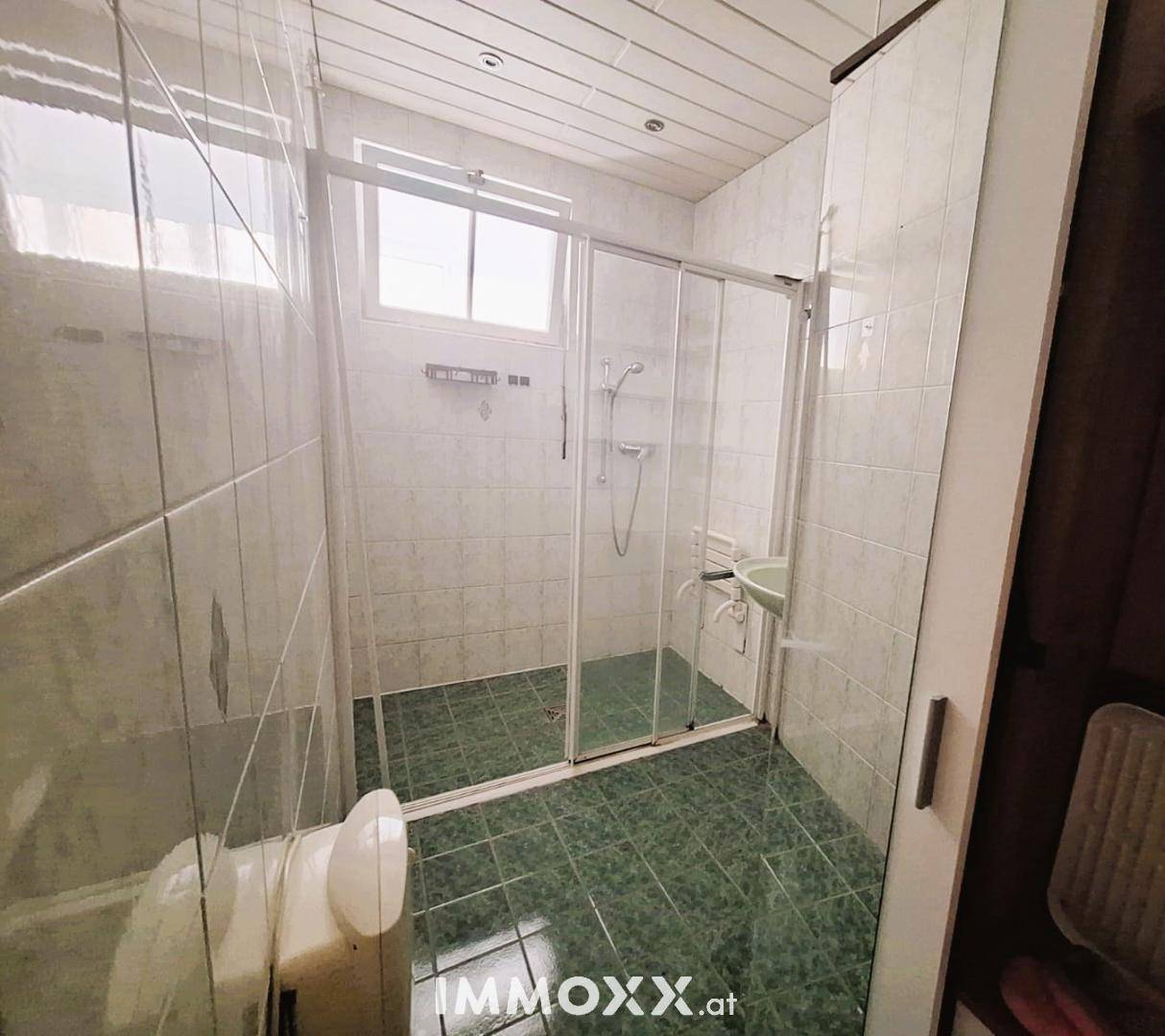 Badezimmer mit grünen Fliesen, Duschkabine und Fenster, das für natürliche Belüftung sorgt.