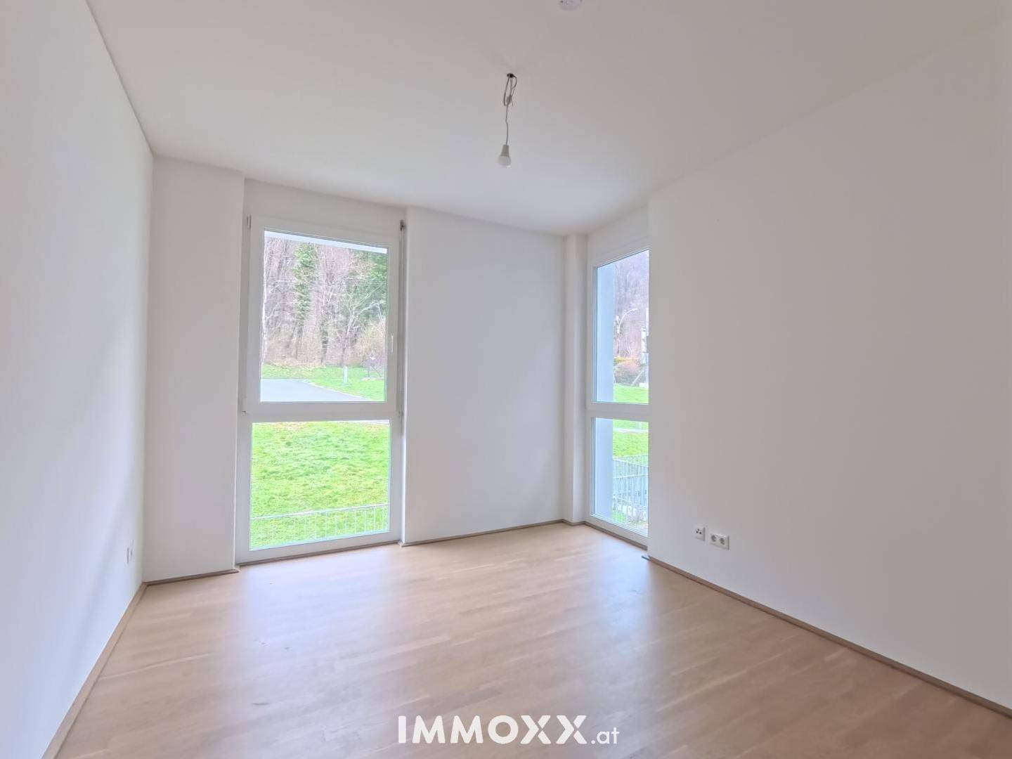 Leerer Raum mit hellem Parkettboden und großen Fenstern, die viel Tageslicht hereinlassen.