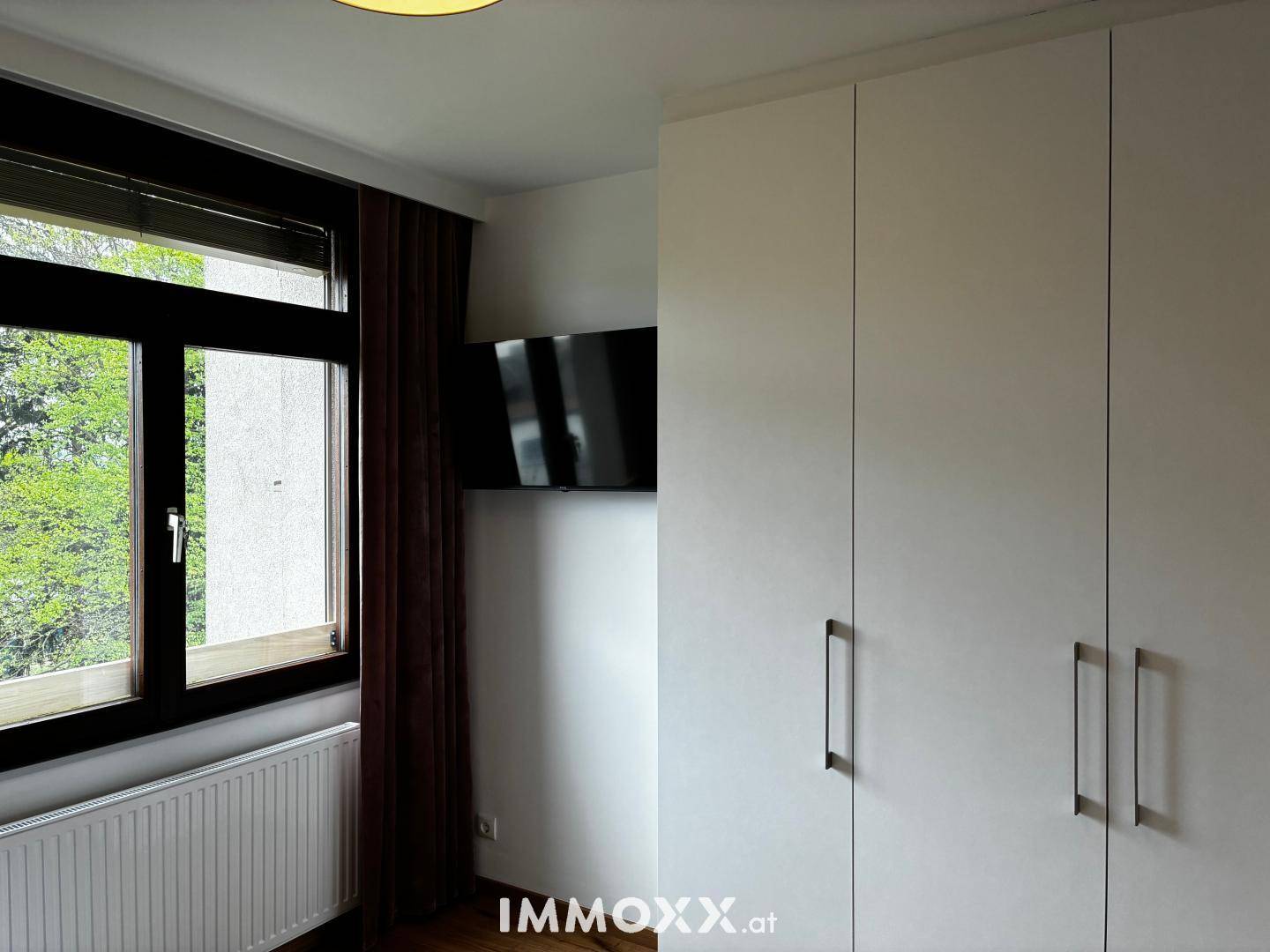 Schlafzimmer mit weißem Kleiderschrank, Wandfernseher und Fenster mit Blick ins Grüne.