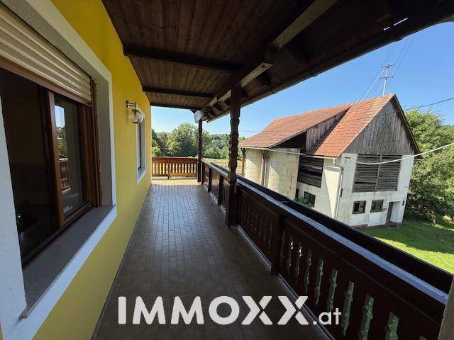 Langer, überdachter Holzbalkon mit traditionellem Geländer und Blick auf ein Nebengebäude und die Landschaft.