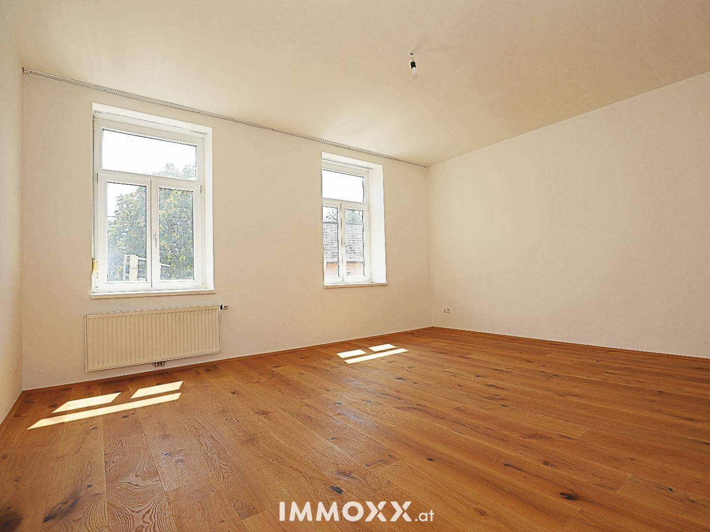 Geräumiger, heller Raum mit Holzboden und zwei Fenstern, die viel Tageslicht hereinlassen.