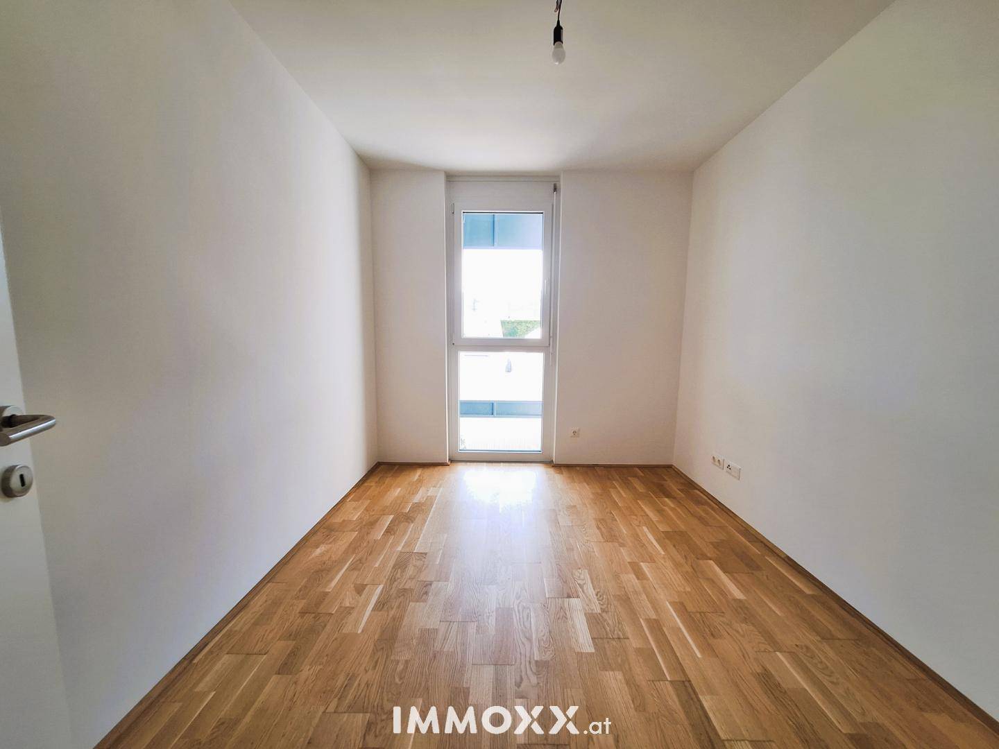 Schlafzimmer mit Parkettboden und großem Fenster, das viel Tageslicht hereinlässt.