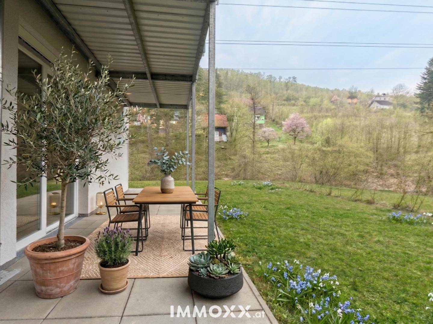 Überdachte Terrasse mit Esstisch und Pflanzen, bietet einen ruhigen Blick ins Grüne.