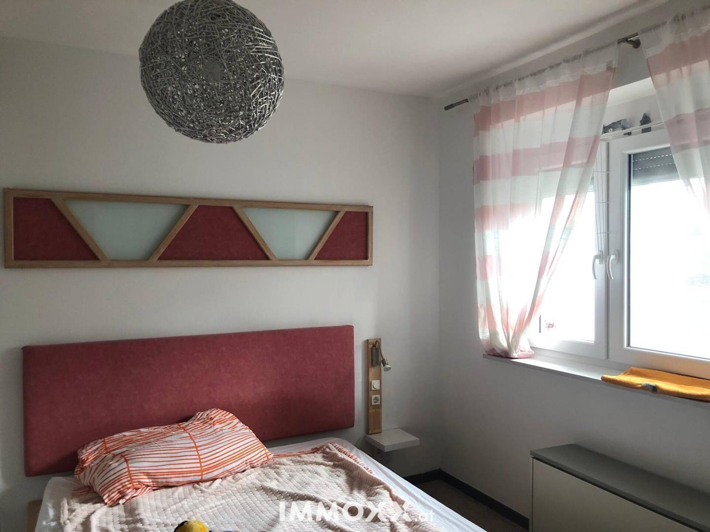 Schlafzimmer mit Einzelbett, rotem Kopfteil und Fenster mit hellen Vorhängen.