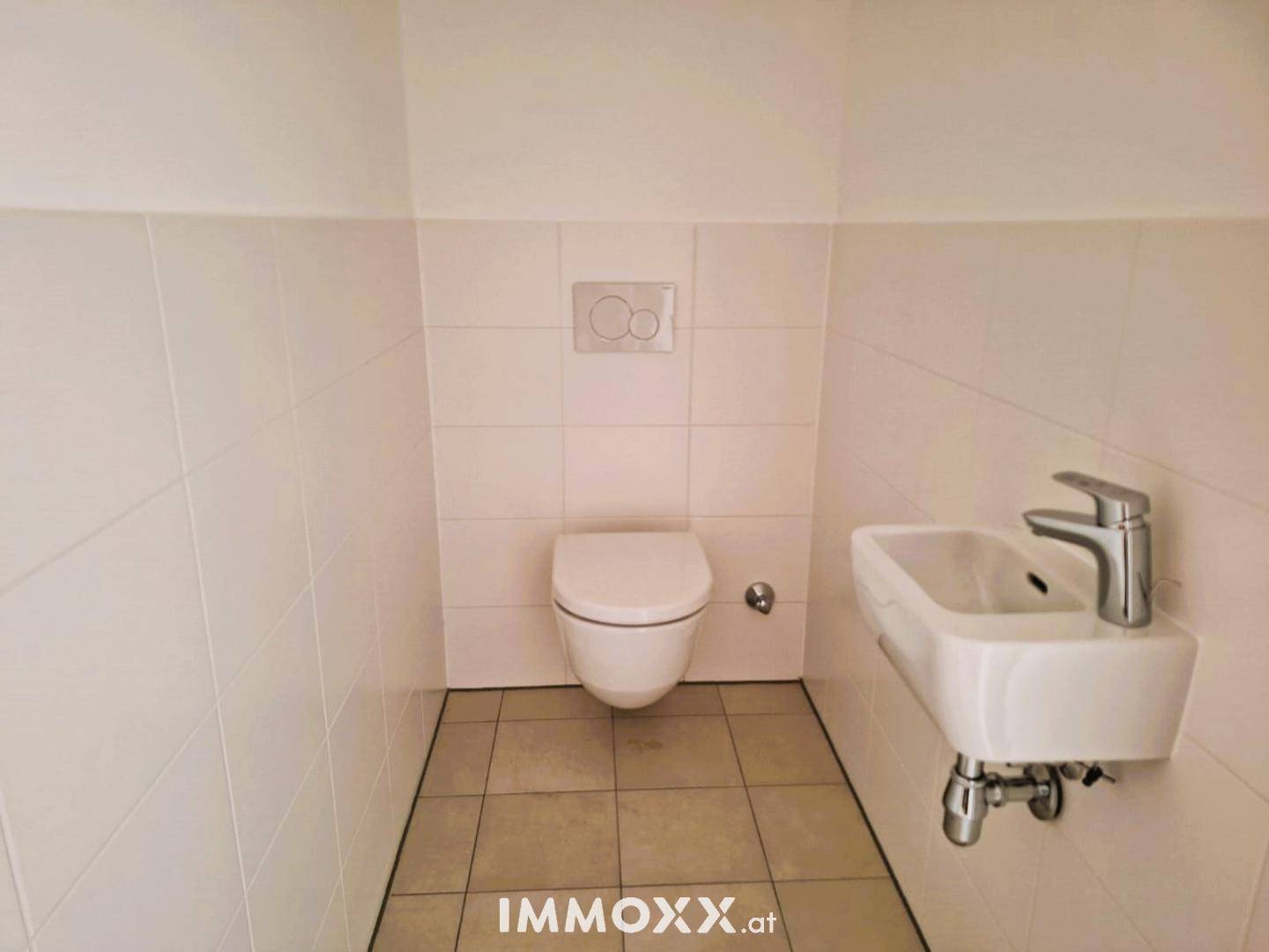 Separates WC mit wandhängender Toilette und kleinem Handwaschbecken, hell gefliest.