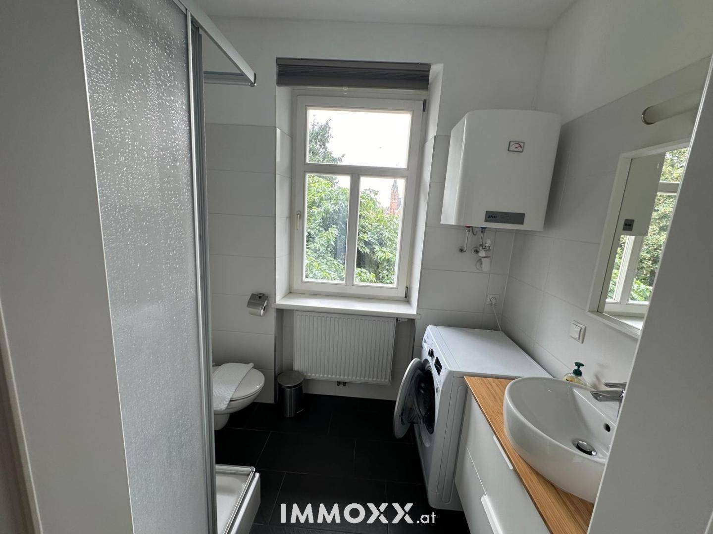 Modernes Badezimmer mit Dusche, WC, Waschmaschine und Fenster mit Blick ins Grüne.