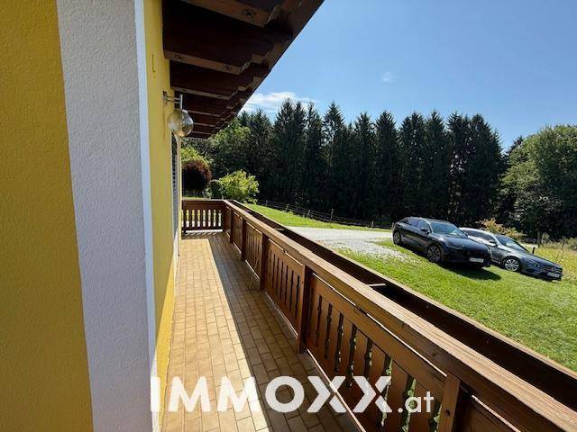 Überdachter Balkon mit Holzboden und Geländer, bietet einen weiten Blick auf die umliegende Natur und Parkplätze.
