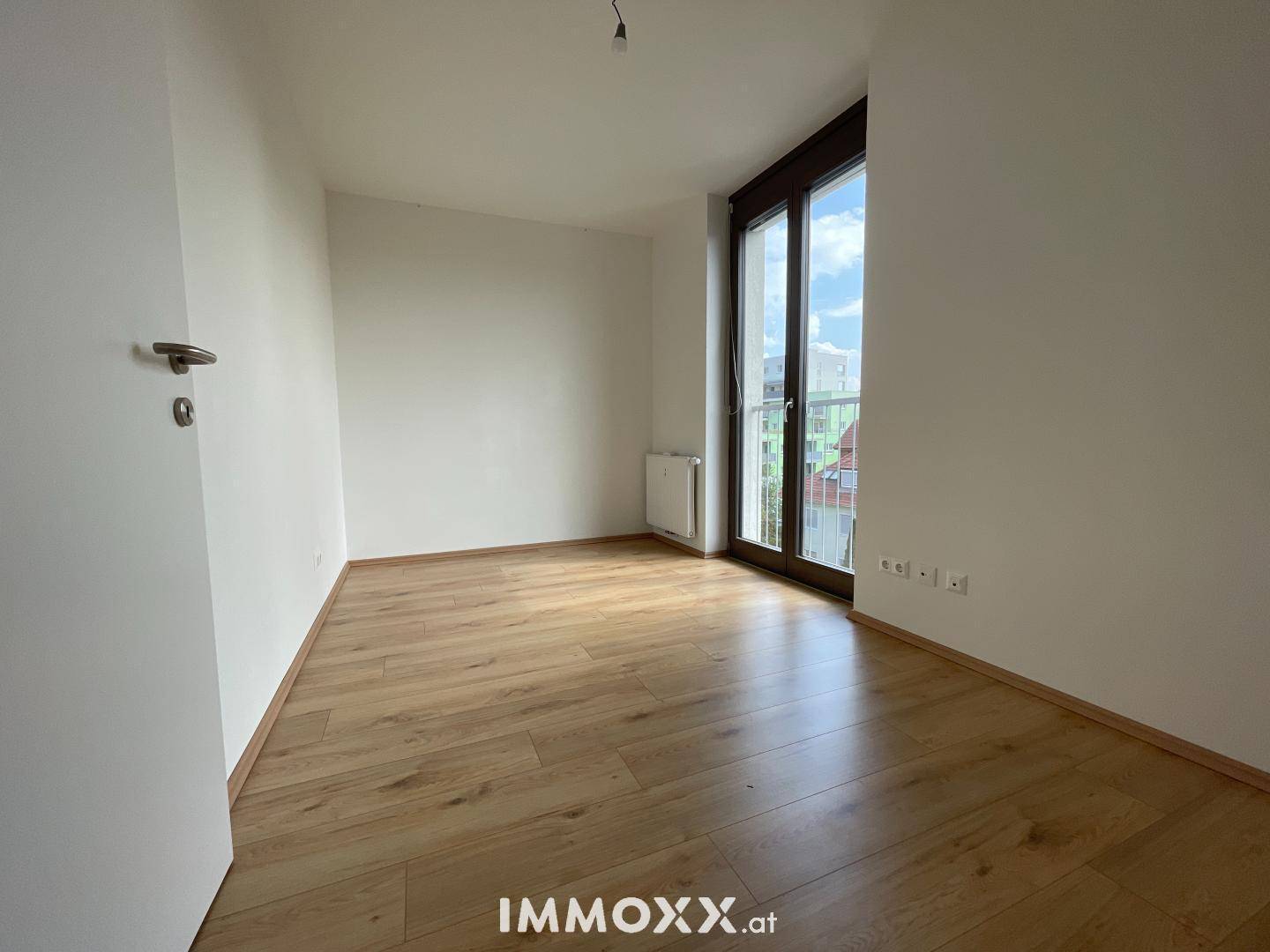 Helles Schlafzimmer mit Laminatboden, Heizkörper und Zugang zu einem französischen Balkon.
