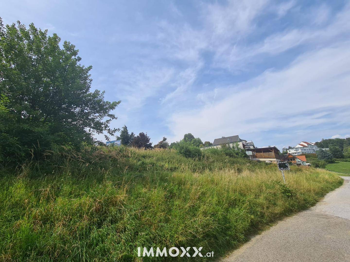 Grünes, hügeliges Grundstück mit hohem Gras und vereinzelten Häusern in der Ferne unter blauem Himmel.