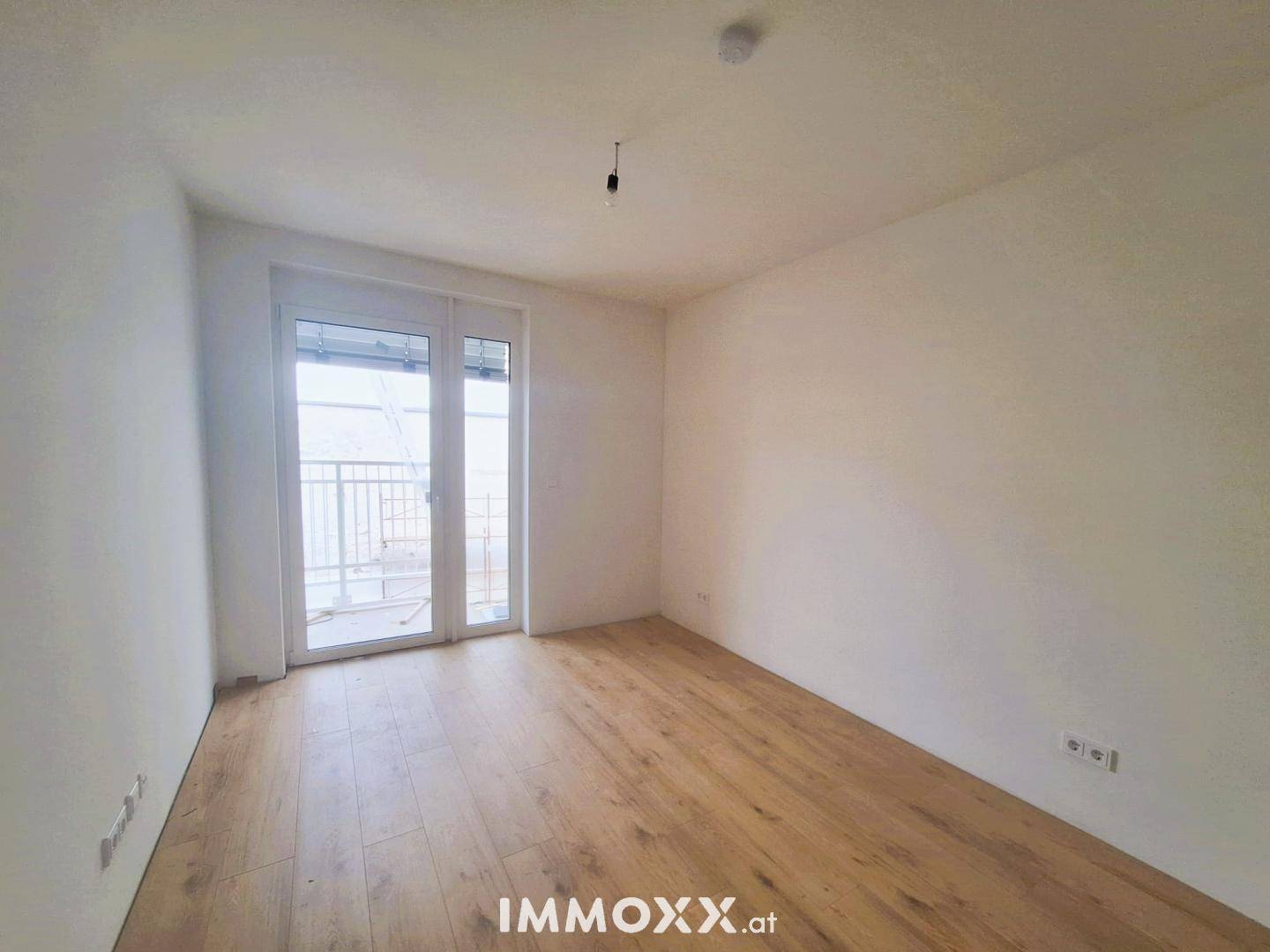 Leeres Zimmer mit Parkettboden und großem Fenster, das viel Tageslicht hereinlässt.