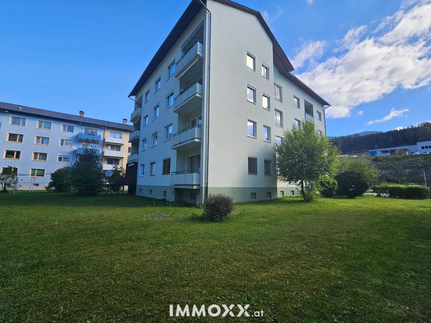Apartmentgebäude mit Balkonen und weitläufiger Rasenfläche vor einer bewaldeten Hügellandschaft.