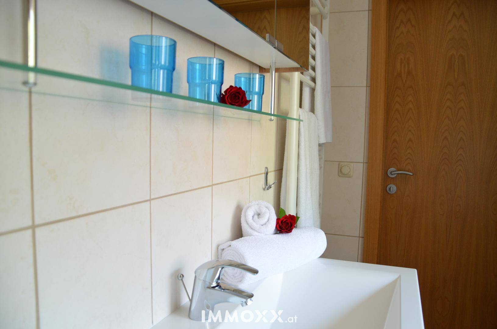 Badezimmer mit modernem Waschbecken, hellen Fliesen und einer Glasablage mit dekorativen blauen Gläsern.