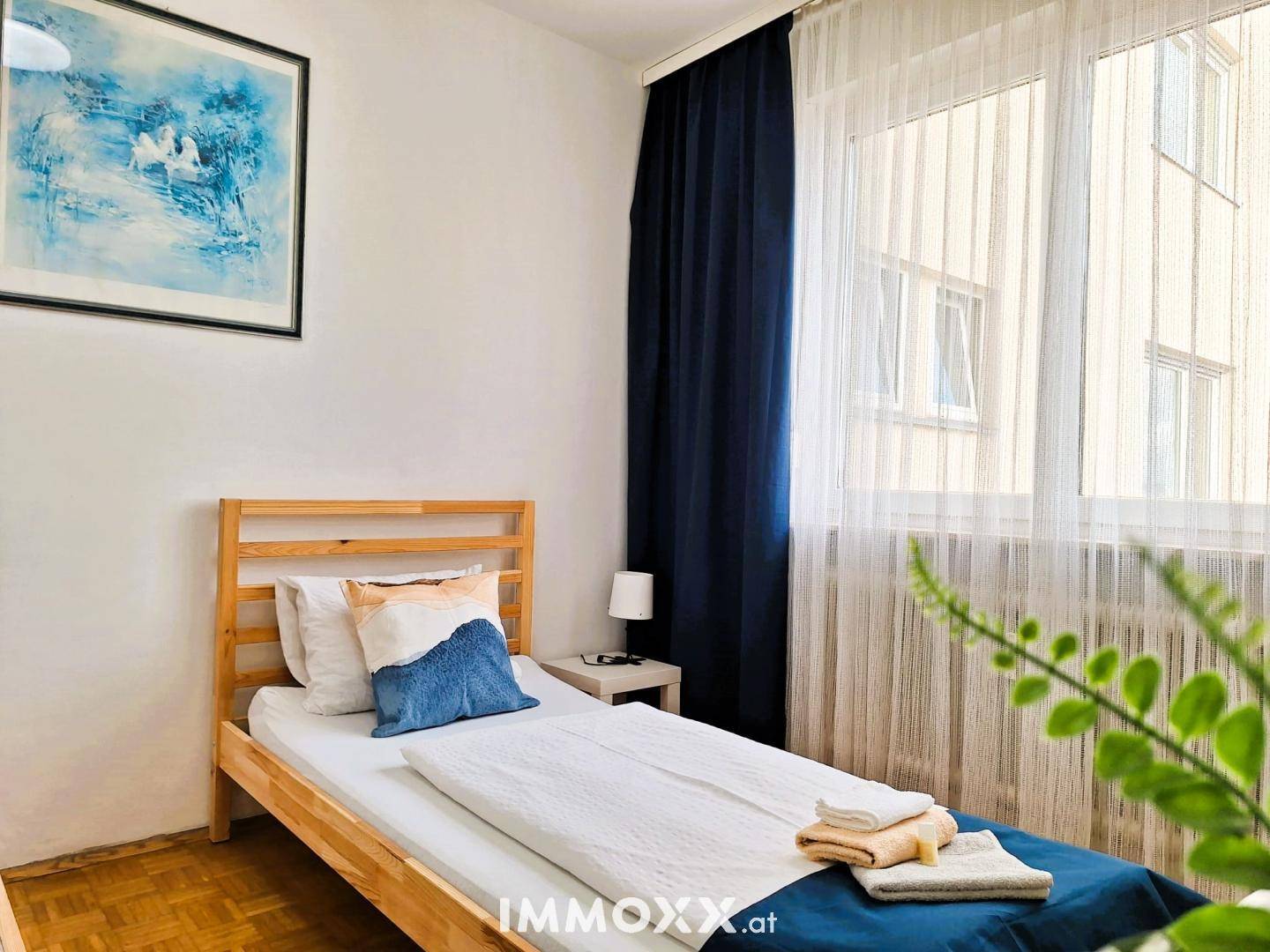 Helles Schlafzimmer mit Einzelbett, Parkettboden und großem Fenster mit blauen Vorhängen.