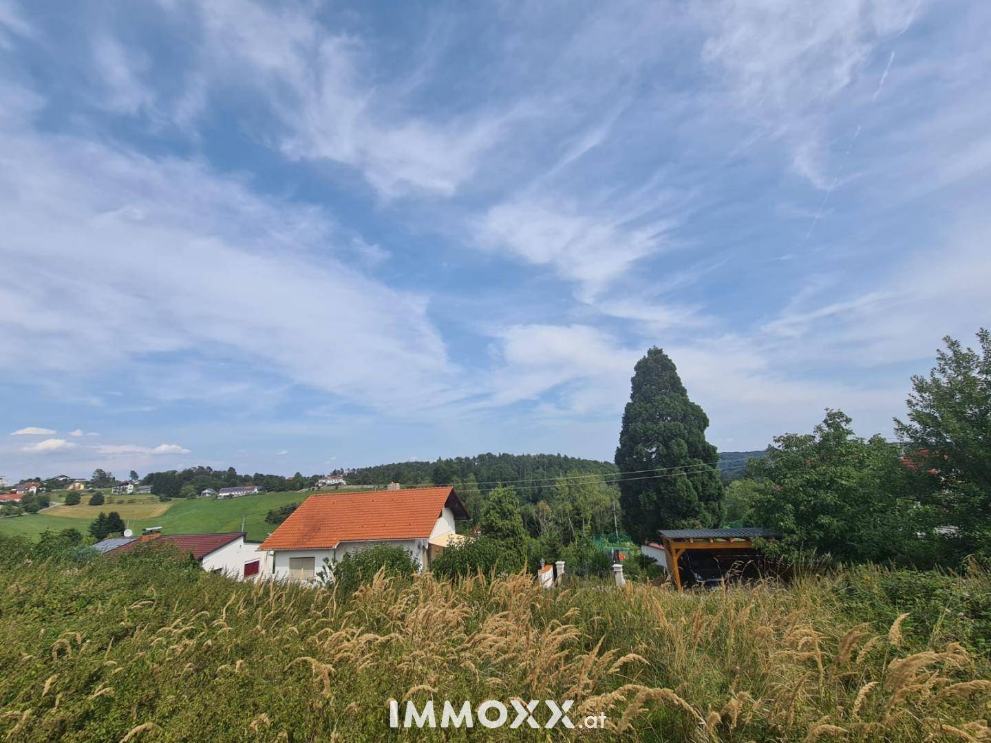 Idyllischer Ausblick über ein Feld auf eine Siedlung mit traditionellen Häusern und bewaldeten Hügeln.