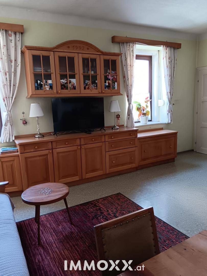 Gemütliches Wohnzimmer mit traditionellen Holzmöbeln, Teppich und Vorhängen an den Fenstern.